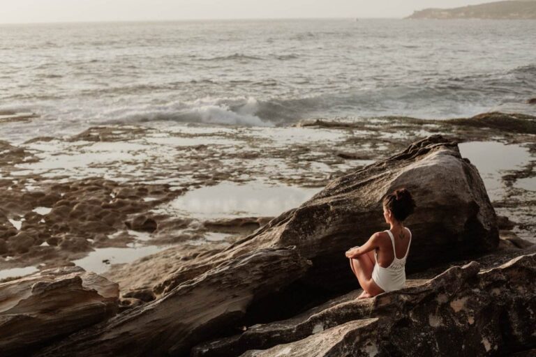 Woman in a white bathing suit sitting sublimely on a rocky ledge happy with herself because she made good choices